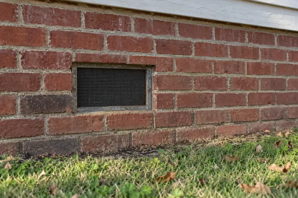 Screened metal vent set low in a faded red brick wall, just above the lawn